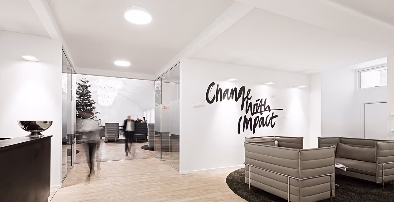 Zona de estar de escritório com sofás cinzentos e parede com a frase Change with Impact sob teto acústico contínuo